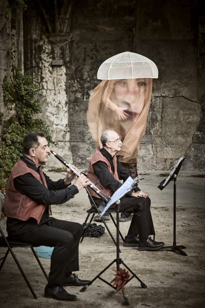 Deux musiciens à l'avant-plan, et la danseuse La Ribot sous un voile imprimé d'un visage.