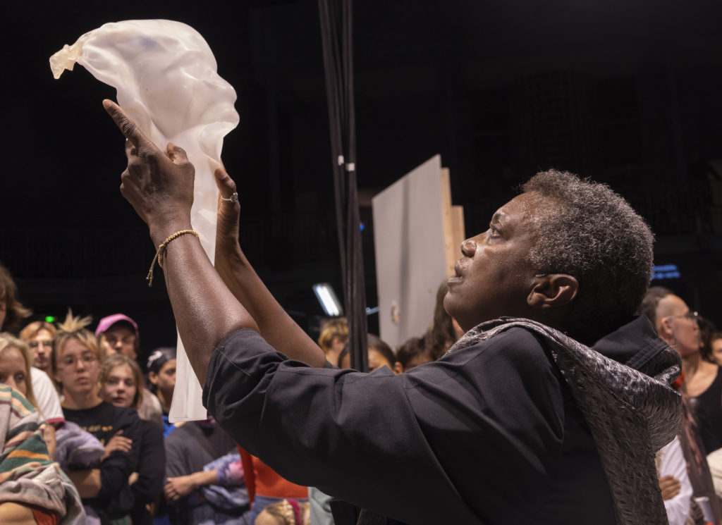 La soprano Claron McFadden, de profil, tenant un moulage d'une partie de corps humain - entourée par le public.